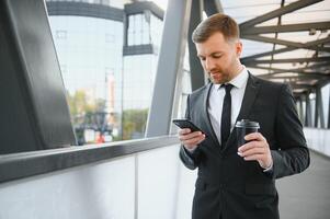 barbado empresario en formal traje en descanso utilizando móvil teléfono utilizar teléfono inteligente negocio hombre en pie fuera de en moderno urbano ciudad calle antecedentes con café taza en céntrico al aire libre. Copiar espacio foto