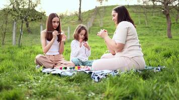 un familia de Tres muchachas de diferente siglos y europeo apariencia son sentado en el jardín en el verde césped y son comprometido en pintura Pascua de Resurrección huevos. Pascua de Resurrección tradiciones primavera día festivo. video